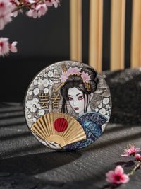 Decorative coaster with a geisha design and cherry blossoms on a dark surface.