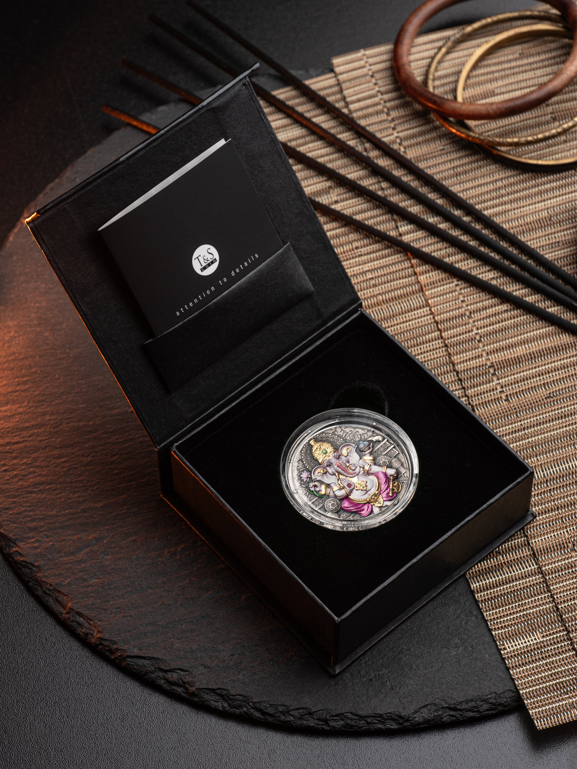 Silver coin with colorful elements in a black box on a dark surface with a woven mat.