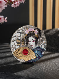 Decorative coaster with a geisha design and cherry blossoms on a dark surface.